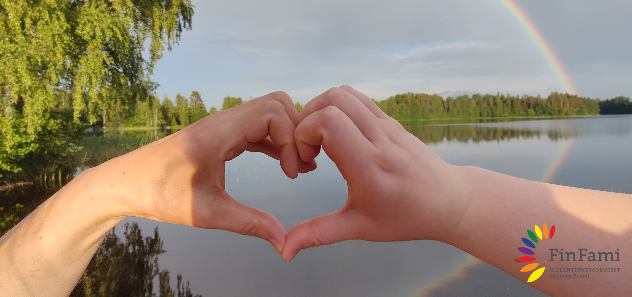Kädet muodostavat sydämen ja taustalla näkyy maisema sekä sateenkaari